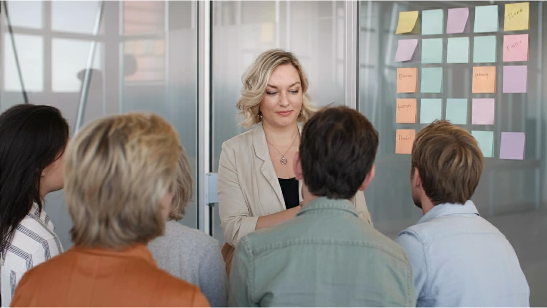 Eine Frau leitet einen Workshop vor einer kleinen Gruppe. Alle blicken aufmerksam auf die Moderation und bunte Notizzettel an der Glaswand. Die Stimmung ist konzentriert, ruhig und gemeinschaftlich.