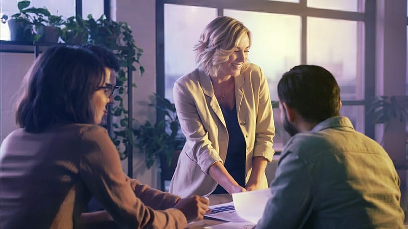 Eine Frau steht an einem Tisch mit zwei Personen und bespricht Unterlagen. Das warme Licht fällt durch große Fenster und vermittelt Fokus, Vertrauen und den gemeinsamen Schritt in die Umsetzung.