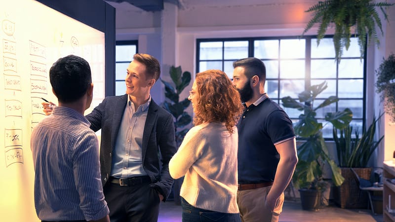 Vier Personen stehen vor einem Whiteboard im hellen Loft, betrachten gemeinsam Notizen und Ideen. Das Bild vermittelt Fokus, Struktur und den Moment, in dem Klarheit entsteht.